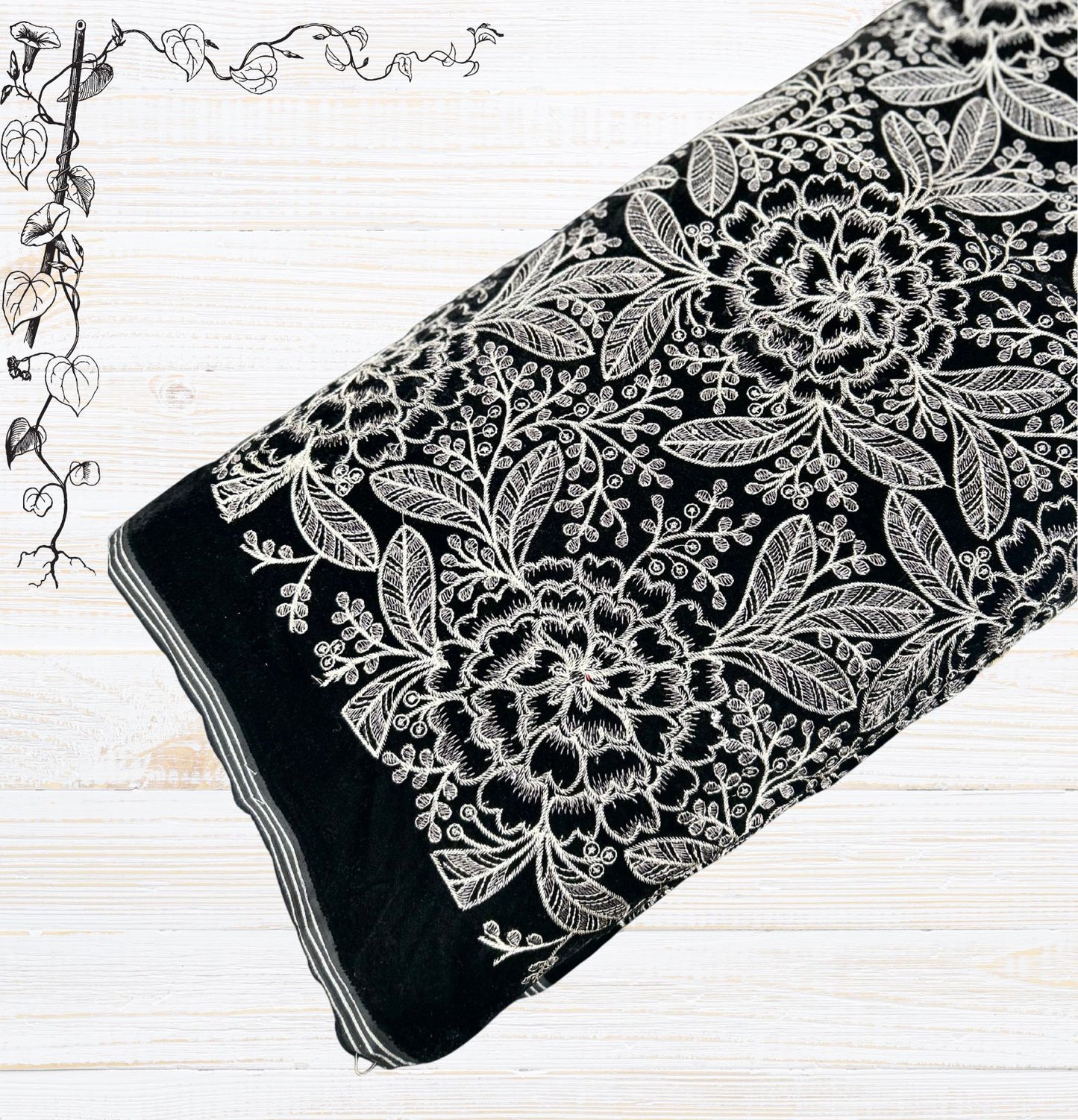 : Luxurious new style premium black velvet fabric with striking white and silver thread embroidery. Features a dense and intricate floral and leafy pattern, perfect for elegant ethnic wear,  formal jackets, or sophisticated home decor. The fabric is rolled, showcasing its plush texture against a light wooden background.