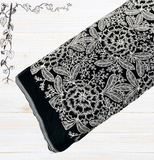: Luxurious new style premium black velvet fabric with striking white and silver thread embroidery. Features a dense and intricate floral and leafy pattern, perfect for elegant ethnic wear,  formal jackets, or sophisticated home decor. The fabric is rolled, showcasing its plush texture against a light wooden background.